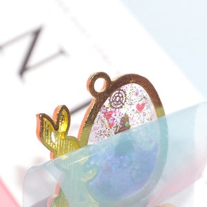 Pode incluir: Encanto de metal dourado com uma resina transparente cheia de glitter e pequenos elementos decorativos. O encanto tem a forma de uma meia lua e tem um pequeno la&ccedil;o na parte superior para prender a um colar ou pulseira.