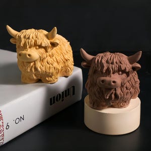 以下が含まれることがあります： ハイランド牛の置物2つ。片方は薄いタン色、もう片方は赤茶色です。どちらも毛むくじゃらの毛皮と角があり、白い本とベージュの台座の上に置かれています。本には「Union」の文字が印刷されています。