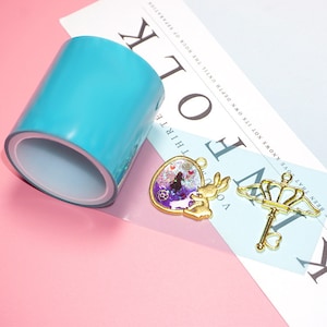 Pode incluir: Um rolo de fita de resina azul transparente, um pingente dourado com um design de resina roxo e azul, e uma chave dourada com uma coroa no topo.