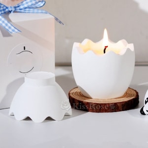 May include: White egg-shaped candle with a lit wick, sitting on a wooden coaster. The candle is in a white egg-shaped container with a cracked top.