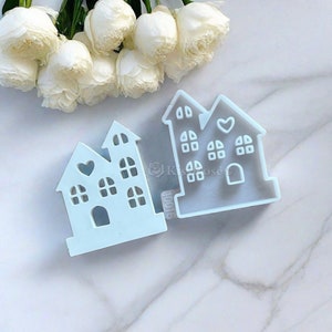 Puede incluir: Dos moldes de silicona azul claro con forma de casas con un detalle en forma de coraz&oacute;n. Los moldes est&aacute;n sobre una superficie de m&aacute;rmol blanco.