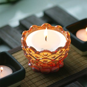 May include: A clear glass candle holder with a decorative, scalloped design. The holder is filled with a white candle that is lit. The candle holder is sitting on a woven mat.