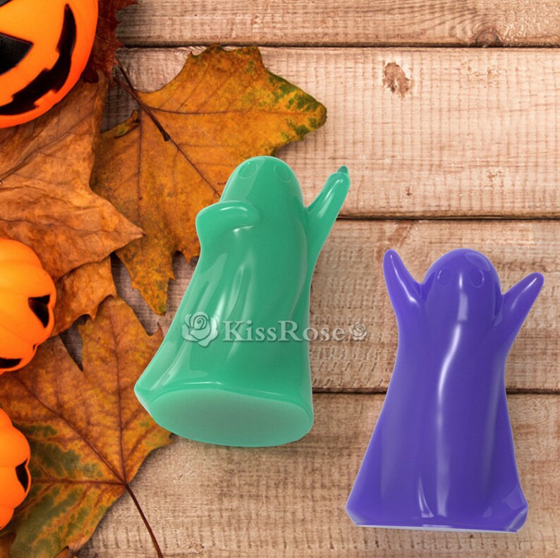 May include: Two Halloween ghost figurines, one green and one purple, on a wooden background. The ghosts are in a standing position with their arms raised.