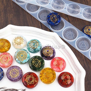 May include: A set of 14 round resin cabochons with gold geometric designs representing the seven chakras. The cabochons are arranged in a circle on a white surface. The cabochons are made with different colored stones, including red, orange, yellow, green, blue, indigo, and violet.