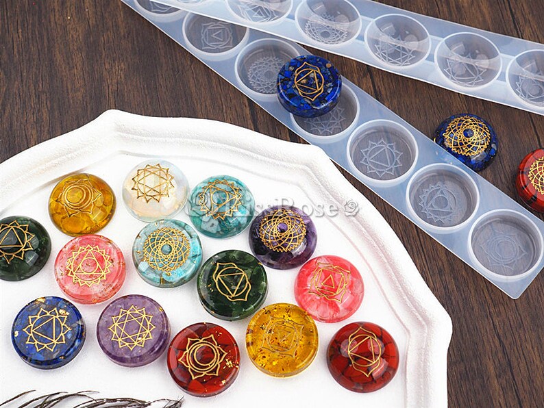 May include: A set of 14 round resin cabochons with gold geometric designs representing the seven chakras. The cabochons are arranged in a circle on a white surface. The cabochons are made with different colored stones, including red, orange, yellow, green, blue, indigo, and violet.