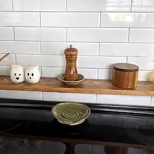 May include: A rectangular wooden stove shelf with a medium brown finish positioned horizontally over a black glass stovetop. The shelf holds a clear glass jar, two white ghost-shaped shakers, a wooden pepper mill on a small plate, and a wooden canister.