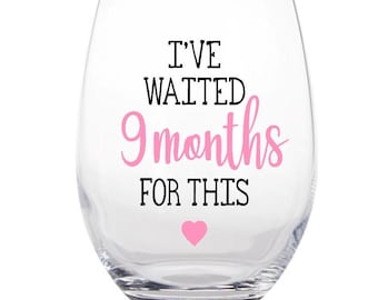 May include: A clear stemless wine glass with the text "I'VE WAITED 9 months FOR THIS" in black and pink lettering. A pink heart is below the text. The glass is designed for a special occasion.