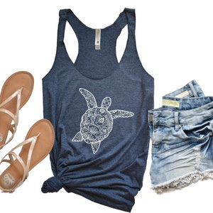 May include: A navy blue tank top with a white graphic of a stylized sea turtle with floral details. The tank top is paired with denim shorts and sandals.