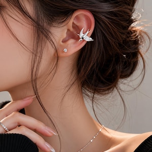 Peut inclure: Un ear cuff argenté en forme d'oiseau, porté sur le haut de l'oreille. Une petite boucle d'oreille ronde est également visible. Le bijou est porté par une personne aux cheveux foncés et un haut noir. Une bague et un collier en argent sont également visibles.