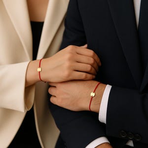May include: Two red string bracelets, each with a small, round gold charm. One bracelet is on a person's wrist wearing a cream-colored blazer, and the other is on a person's wrist wearing a navy suit jacket.