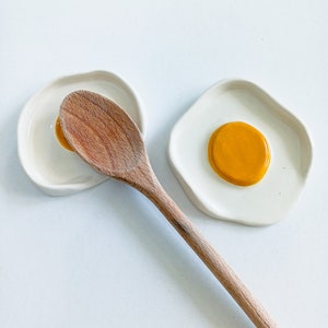 Op de afbeelding: Twee witte keramische lepelsteunen in de vorm van gebakken eieren, één met een houten lepel erop. De lepelsteunen hebben een gele dooier in het midden. De houten lepel is lichtbruin.