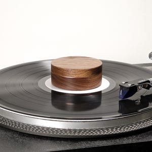 May include: A circular dark brown wooden record weight with a smooth grain finish, positioned centrally on a black vinyl record resting on a turntable platter. The record player features a silver tone arm with a blue and black cartridge attached.