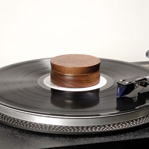 May include: A circular dark brown wooden record weight with a smooth grain finish sits centered on a black vinyl record on a turntable. The record player features a silver platter edge and a black tonearm with a blue Ortofon cartridge.