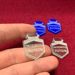 Puede incluir: Cuatro fichas de poción de vidrio transparente con una inscripción blanca que dice "Poción de invisibilidad". Dos de las fichas son azules y dos son transparentes.