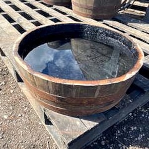 May include: A large, circular wooden barrel filled with water, reflecting the sky. The barrel has a dark, weathered appearance with metal bands. It sits on a wooden pallet, with two other barrels visible in the background. The water's surface is calm.