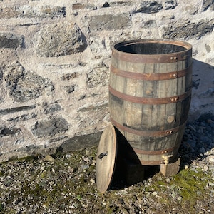 May include: A large wooden barrel with metal bands and a lid. The barrel is sitting on a stone surface and is surrounded by gravel.