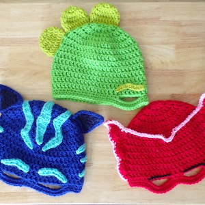 May include: Three handmade crochet hats in bright colors. A green dinosaur hat with yellow accents, a blue and turquoise cat-like hat, and a red mask-style hat with pink trim. All hats are made with a visible crochet stitch.