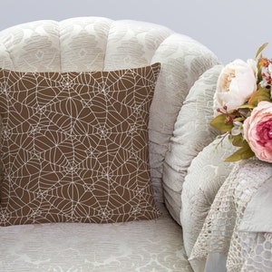 May include: A brown throw pillow with a white spiderweb pattern. The pillow is sitting on a white armchair with a white crocheted throw draped over the arm.