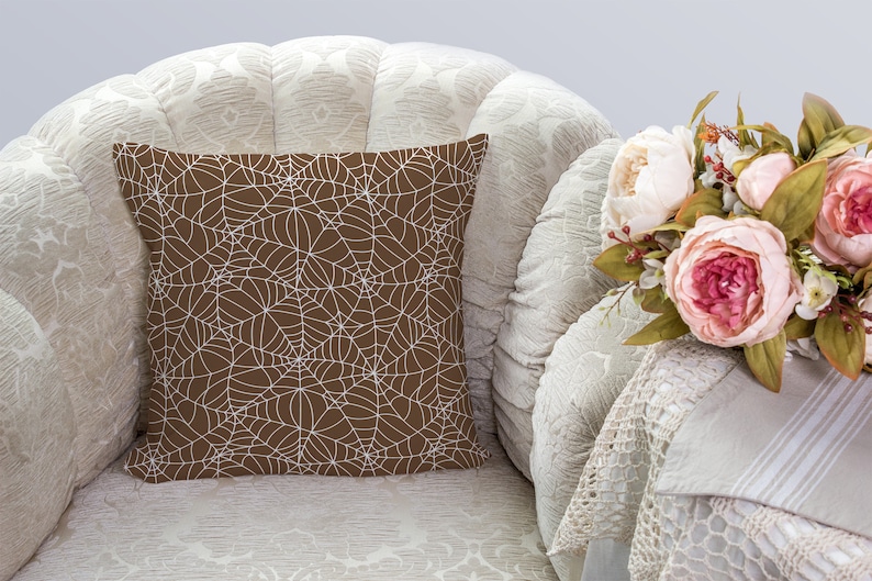 May include: A brown throw pillow with a white spiderweb pattern. The pillow is sitting on a white armchair with a white crocheted throw draped over the arm.