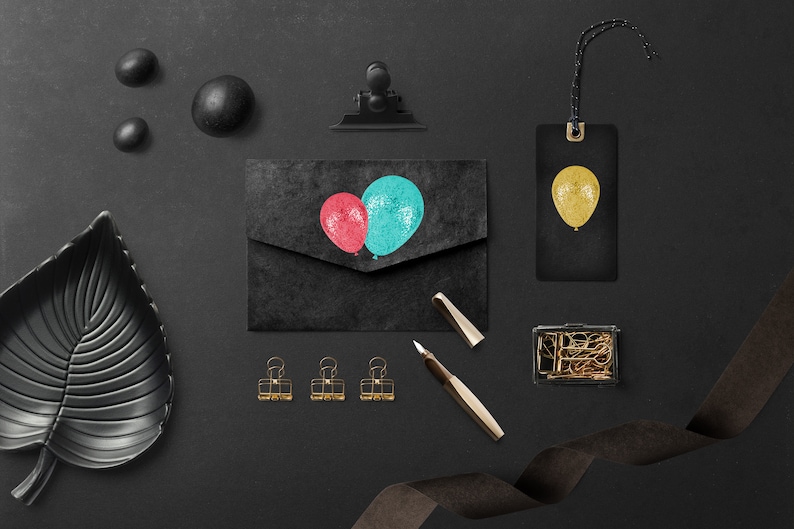 May include: A black envelope with a red and blue balloon design. The envelope is on a black surface with a gold ribbon, a gold box, and a black leaf-shaped dish.