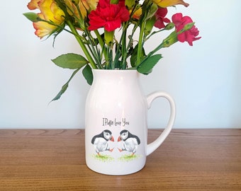 May include: White ceramic pitcher with a handle, featuring a painted design of two puffins with the text "I Puffin Love You". The pitcher is filled with a bouquet of pink and yellow carnations.