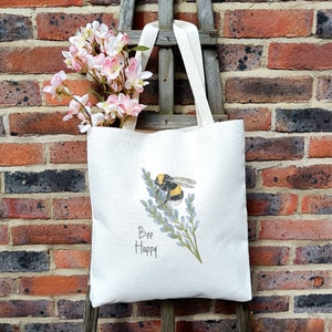May include: A white tote bag with a bee and lavender flowers printed on it. The text "Bee Happy" is printed below the image. The bag is sitting on a brick wall with pink flowers in the background.