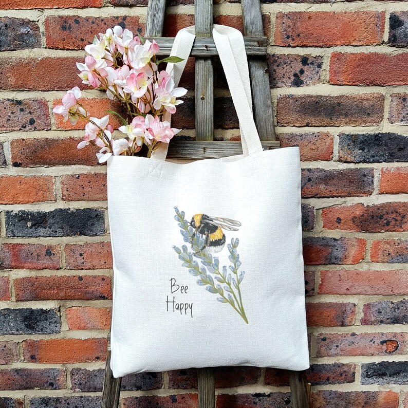 May include: A white tote bag with a bee and lavender flowers printed on it. The text "Bee Happy" is printed below the image. The bag is sitting on a brick wall with pink flowers in the background.