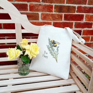 May include: A white tote bag with a bee and flowers design. The bag has a black strap and the text "Bee Happy" printed on it.