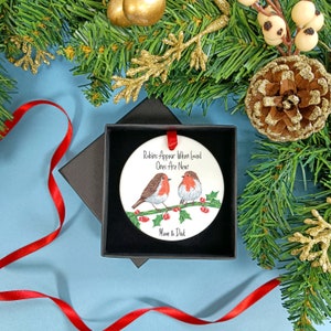 May include: White ceramic Christmas bauble with a red ribbon. The bauble features a painting of two robins perched on a holly branch with red berries. The text on the bauble reads "Robins Appear When Loved Ones Are Near" and "Mum & Dad".
