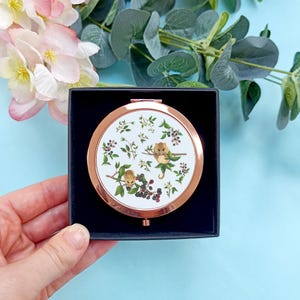 May include: A rose gold compact mirror with a white background and a design of two brown and white dormice on a branch with green leaves and black berries.