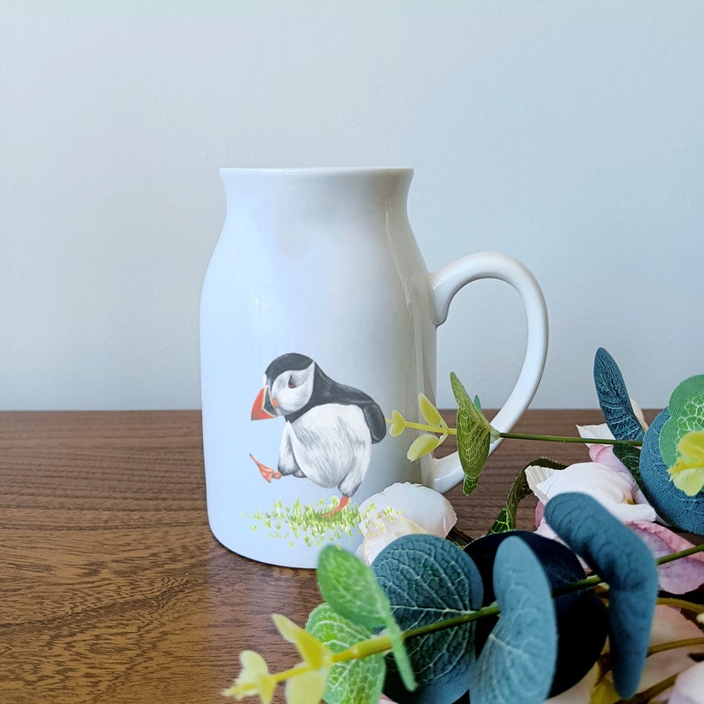 Op de afbeelding: Een witte keramische mok met een handvat, met een handgeschilderde illustratie van een papegaaiduiker met een zwart-wit patroon en een oranje snavel. De papegaaiduiker staat op een groene grasveld. De mok staat op een houten oppervlak met groene bladeren op de voorgrond.