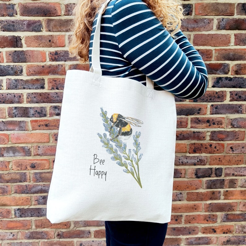 May include: A white canvas tote bag with a bee and lavender flowers printed on it. The text "Bee Happy" is printed below the bee.