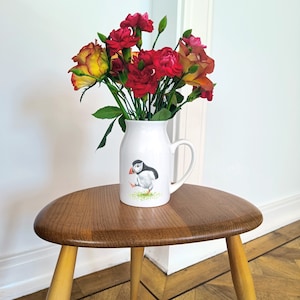 Op de afbeelding: Een witte keramische kan met een puffin-illustratie, gevuld met een boeket rode en gele bloemen. De kan heeft een handvat en staat op een houten tafel.