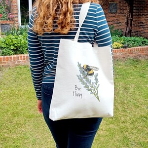 May include: White tote bag with a bee and lavender flowers printed on it. The text "Bee Happy" is printed below the bee.