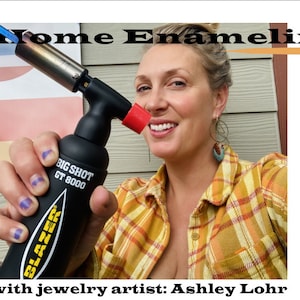 May include: A person holding a black torch with the words "BIG SHOT GT 8000" and "BLAZER" printed on it. The torch is lit and has a blue flame. The person is wearing a yellow and orange plaid shirt and has a turquoise earring.