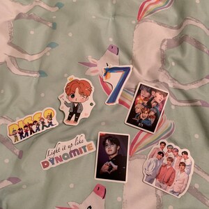 May include: A collection of seven stickers featuring the South Korean boy band BTS. The stickers include a cartoon drawing of a band member, a group photo of the band, a photo of a band member making a heart shape with his hands, and a sticker with the text "Light it up like Dynamite".