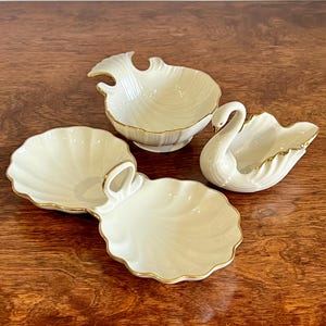 May include: Four ivory porcelain serving dishes with gold edging. The set includes a swan-shaped dish, a shell-shaped dish, and two other dishes with scalloped edges. The dishes are displayed on a wooden surface.