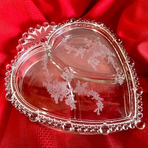 Puede incluir: Un plato de servir de vidrio transparente en forma de corazón con tres secciones. El plato tiene un borde con cuentas y un delicado patrón floral grabado en el vidrio.