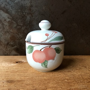 Puede incluir: Un azucarero de cerámica blanca con tapa, con un diseño floral con manzanas rosas y hojas verdes. El tazón tiene un borde marrón y un pequeño pico para verter.