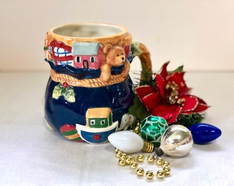 Pode incluir: Uma caneca de Natal de cerâmica com base azul marinho e borda bege, decorada com um urso de pelúcia, casas e um barco. A caneca é cercada por enfeites de Natal, incluindo bolas e contas douradas.
