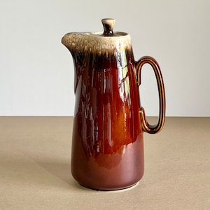 Może przedstawiać: Wysoki, brązowy ceramiczny dzbanek z pokrywką. Dzbanek ma uchwyt i dzióbek. Górna część dzbanka jest jaśniejsza, z efektem spływającej glazury. Dzbanek jest prawdopodobnie przeznaczony do serwowania napojów.