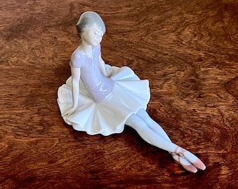 May include: A porcelain figurine of a ballerina wearing a white tutu and a lavender leotard. She is sitting with her legs extended and her head tilted down.