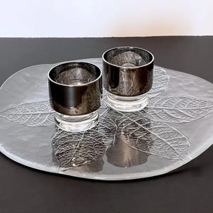 May include: Two silver-rimmed glass candle holders sit on a clear, leaf-patterned glass tray. The candle holders have a clear base and a dark, metallic finish. The tray is oval-shaped with a frosted leaf design.