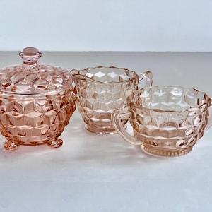 Op de afbeelding: Een roze glazen serveerset van drie delen. De set bevat een suikerpot met deksel, een melkkan en een suikerpot met handvatten. Elk stuk heeft een geometrisch, diamantachtig patroon.