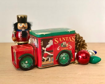 May include: A red Santa's Express truck tin with green wheels and a green top, featuring Santa Claus inside. A wooden nutcracker stands beside the truck. Christmas ornaments and a pinecone are also present.