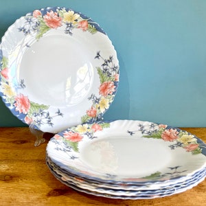 Puede incluir: Un juego de platos de cerámica blanca con borde festoneado azul y diseño floral. Los platos presentan flores rosas, amarillas y azules. Los platos están apilados sobre una superficie de madera.