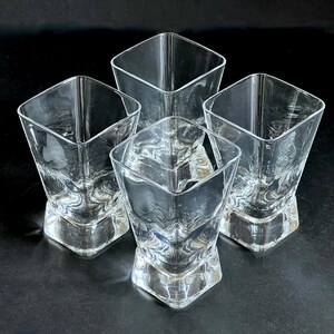 May include: Four clear, square-shaped drinking glasses. Each glass has a textured base and a flared, angled top. The glasses are made of transparent glass and are arranged on a dark surface, creating a play of light and shadow.