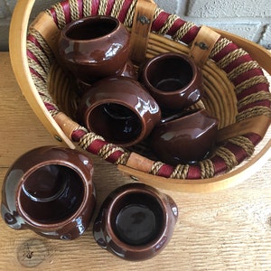 Könnte beinhalten: Ein Set aus sechs braunen Keramiktöpfen in einem geflochtenen Korb mit einem Holzgriff. Die Töpfe sind alle in verschiedenen Größen und Formen und in zufälliger Reihenfolge angeordnet.