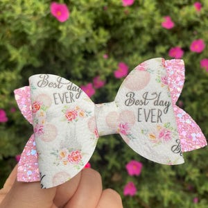 May include: A white and pink hair bow with the text "Best day EVER". The bow features a floral pattern with pink glitter accents on the tips. The background is a blurred view of pink flowers and green foliage.