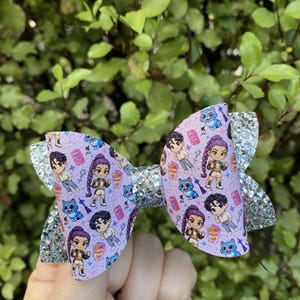 May include: A lavender hair bow with a glittery silver accent. The bow features a repeating pattern of cartoon characters and other small illustrations. The bow is held in front of a green leafy background.